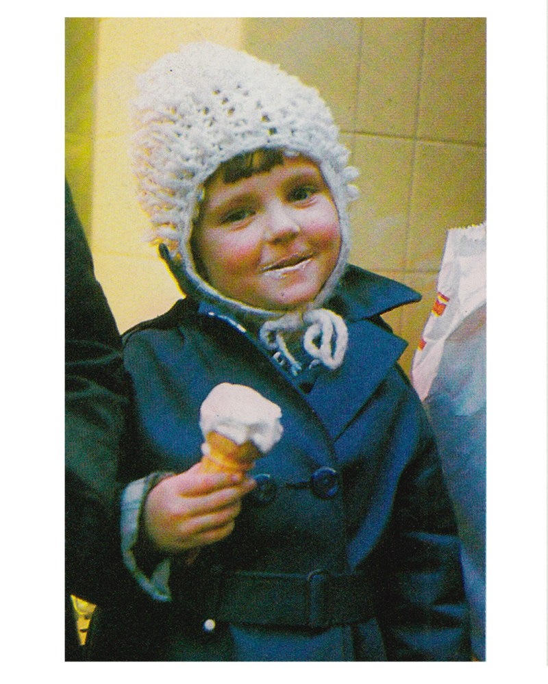 A young child wearing a white knitted hat and a dark coat holds an ice cream cone, smiling at the camera.